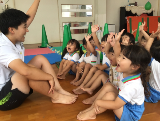 のびのびキッススクールの様子の写真、先生と子供が楽しそうに手を挙げています。