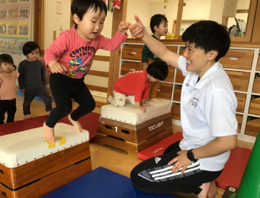 のびのびキッススクールの様子の写真、跳び箱の上からジャンプする子供の手を先生が持ってサポートしています。